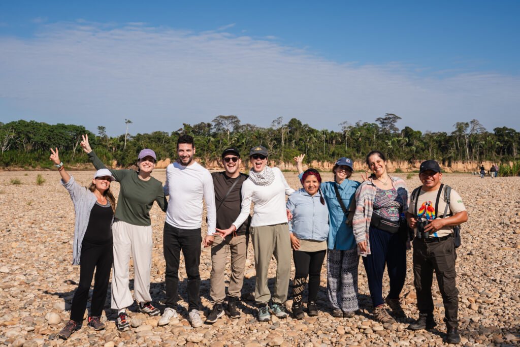A group of smiling travelers hugging on the riverbank during a tour of the macaw clay lick in Tambopata, with the macaw clay cliffs visible in the background.